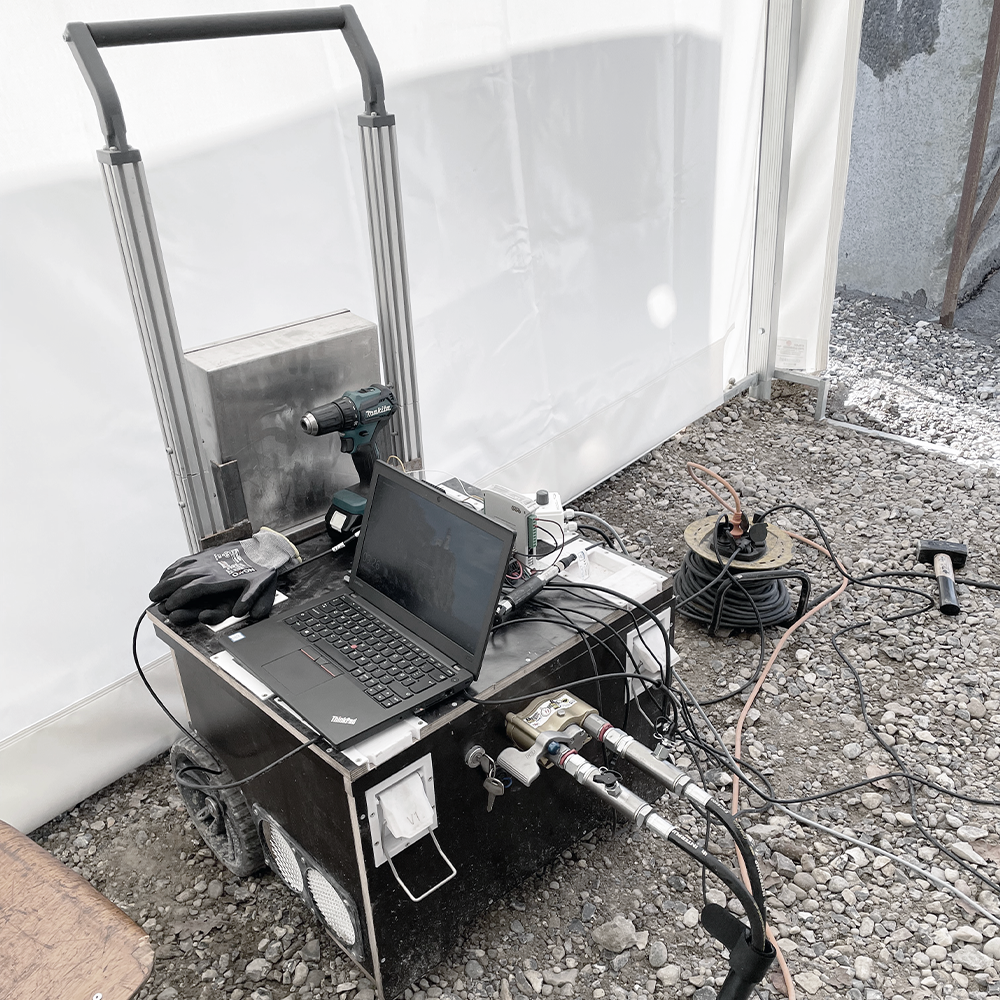 Laptop, arbejdshandsker, boremaskine og ledninger oven på en trækasse monteret på sækkevogn med hydraulikslanger tilkoblet i fronten.